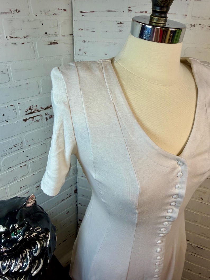 May include: A cream-colored dress with a V-neckline and short sleeves. The dress features a row of small buttons down the front and vertical seams for a tailored look. The dress is displayed on a mannequin.