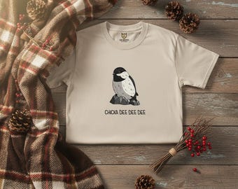 May include: Cream-colored t-shirt featuring a black and white bird illustration perched on a rock. The text "CHICKA DEE DEE DEE" is printed below the bird. The shirt is displayed on a wooden surface with a plaid blanket and pine cones.