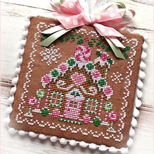 Puede incluir: Un adorno de punto de cruz de una casa de jengibre marrón con detalles de caramelo rosa y blanco. El adorno está decorado con un borde de pompón blanco y una cinta de lunares rosa y blanco.
