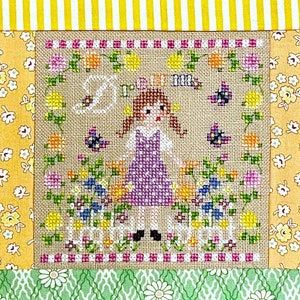 Pode incluir: Bordado de ponto cruz com uma menina em um vestido roxo em pé em um jardim com flores e borboletas. O texto "Good Morning" está bordado acima da menina.