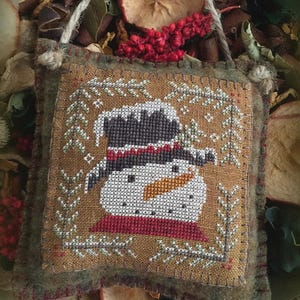 May include: A square, hand-stitched fabric ornament featuring a cross-stitch design of a snowman wearing a black top hat and a red scarf. The snowman is centered on a tan background, framed by stitched pine branches, with a jute hanging loop.