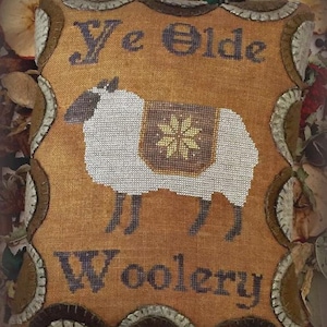 May include: A rectangular fabric pincushion with a mustard-coloured linen front featuring a cross-stitched white sheep with a brown blanket and a yellow star. The text "Ye Olde Woolery" is stitched in dark blue. The edges have a scalloped felt border.