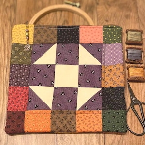 May include: A square patchwork quilt block featuring a central star pattern in cream and purple floral fabric, surrounded by a border of multi-colored floral squares. Includes a wooden embroidery hoop, three spools of thread, and metal scissors.
