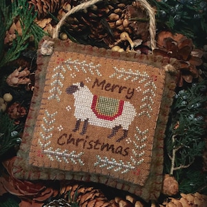 May include: A square, brown fabric ornament featuring a cross-stitched design of a white sheep with a green and red saddle. The text Merry Christmas is stitched in brown. The ornament has a frayed edge and a jute twine loop for hanging.