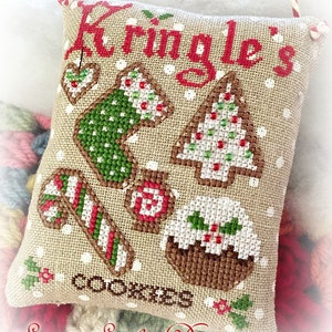 May include: A beige fabric cushion with a white background and red and green embroidered Christmas cookies. The cushion has the text "Kringle's Cookies" and "Sugar Stitches Designs" embroidered on it.