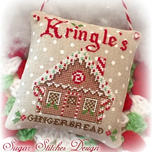 Può includere: Un cuscino beige con pois bianchi presenta un ricamo a punto croce di una casetta di pan di zenzero con dettagli di bastoncini di zucchero. Il cuscino è decorato con il testo "Kringle's Gingerbread" ed è un ornamento natalizio fatto a mano.
