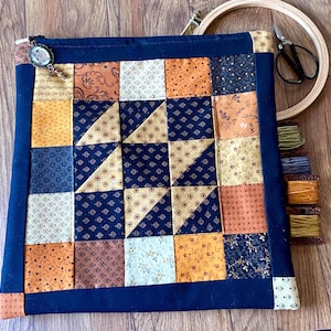 May include: A square quilted project bag featuring a patchwork design of navy blue, orange, and cream fabric squares with floral and geometric patterns. The bag has a navy blue border, a zipper closure with a decorative charm, and embroidery tools.