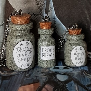 May include: Three small, textured glass bottles with cork stoppers and silver chains. Each bottle has a white label with black text: "Deadly Night Shade", "Frog's Breath", and "Worm's Wort". The background features a character from The Nightmare Before Christmas.