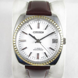 Könnte beinhalten: Eine silberne Citizen Armbanduhr mit weißem Zifferblatt, goldenen Akzenten und einem braunen Lederarmband. Das Zifferblatt der Uhr verfügt über schwarze Zeiger, schwarze Stundenmarkierungen und den Text "17 JEWELS SHOCKPROOF JAPAN MADE".