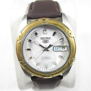Peut inclure: Une montre-bracelet automatique Seiko 5 avec un cadran argenté, une lunette dorée et un bracelet en cuir marron. Le cadran affiche le jour et la date, avec le jour indiquant "SAT 8". La montre est marquée "AUTOMATIC 21 JEWELS" et "MADE IN JAPAN".