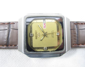 May include: A gold-toned Seiko watch with a square face and a brown leather band. The watch face has a day of the week display, a date window, and the text "Automatic 23 Jewels".