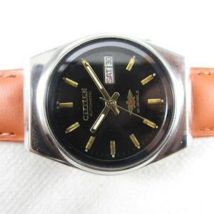 May include: A black-faced Citizen automatic wristwatch with gold-tone hands and hour markers. The watch has a day and date window at the 3 o'clock position and a brown leather band.