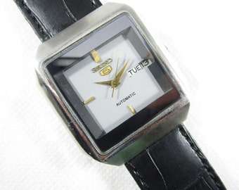 May include: A silver and black square-shaped wristwatch with a white face. The watch has a black leather band and says "SEIKO 5 AUTOMATIC" and "TUE 13" on the face.