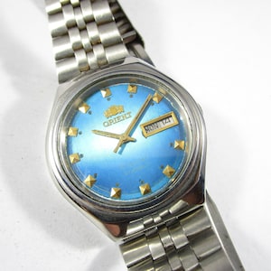 Può includere: Un orologio Orient argentato con quadrante blu e dettagli dorati. L'orologio ha una finestrella per la data che mostra "MON 14".
