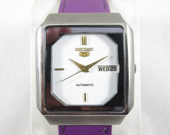 May include: A silver square-shaped wristwatch with a white face, black hands, and a purple leather band. The watch face has the words "SEIKO 5 AUTOMATIC" and the day of the week "WED 25" displayed.