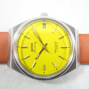 Op de afbeelding: Een geelgekleurd polshorloge met een zilveren band en een zwarte leren band. De wijzerplaat van het horloge heeft zwarte markeringen en de tekst "RHYMA Tarveq PARA SHOCK 17 JEWELS".