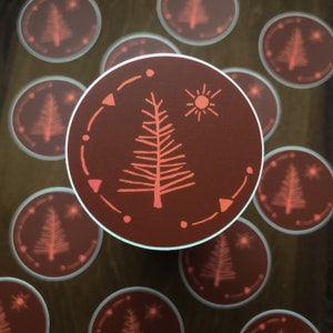 May include: A set of round stickers with a brown background and a pink stylized tree design. The tree is surrounded by a circle of pink dots and triangles. A pink sun is in the upper right corner of the sticker.