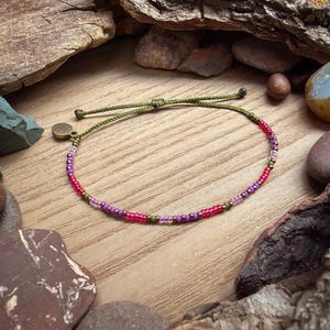 May include: A beaded bracelet with a green cord and a mix of pink, purple, and gold beads. The bracelet is set against a wooden surface with rocks and wood pieces in the background. The bracelet is approximately 7 inches in length.