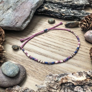 May include: A delicate pink and purple beaded bracelet with a small metal charm. The bracelet is made with tiny beads and has a simple, elegant design.