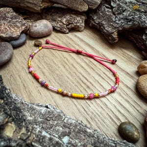 May include: A colorful beaded bracelet with pink, yellow, and red beads, and a gold clasp. The bracelet is on a wooden surface.