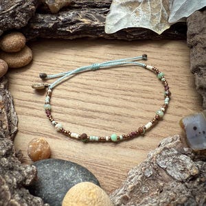 May include: A beaded bracelet with a light blue cord. The bracelet features a mix of small, colorful beads in shades of green, brown, white, and gold. The bracelet is displayed on a wooden surface with natural elements.