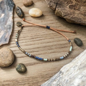 May include: A delicate beaded bracelet with a mix of blue, brown, and white beads. The bracelet is tied with a brown cord and has a small metal clasp.