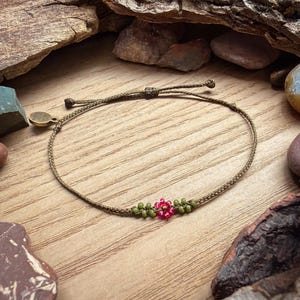 May include: A handmade olive green cord bracelet with a central floral design. The flower is made of small red and green beads. The bracelet has an adjustable sliding knot closure and a small, round, gold-colored charm.