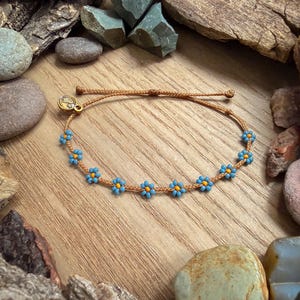 May include: A handmade bracelet featuring a series of small blue and yellow beaded flowers strung on a brown cord. The bracelet has a gold-colored charm and is displayed on a wooden surface.