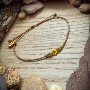 May include: A handmade bracelet featuring a woven brown cord with a small flower design made of yellow and green beads. The bracelet has a gold-colored charm and is displayed on a wooden surface with stones and bark.