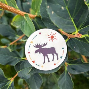 May include: A round sticker featuring a purple moose silhouette, a red sun, and colorful geometric shapes. The moose is centered on a cream-colored background, encircled by a dotted line and decorative elements. The sticker is set against a backdrop of green leaves.