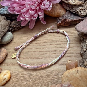 May include: A braided bracelet in pastel colors of pink, yellow, and blue. The bracelet has a small gold-colored charm. A large pink flower and various rocks and wood pieces surround the bracelet on a wooden surface.