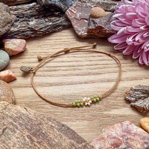 May include: A tan cord bracelet with a daisy design made of white, orange, and green beads. The bracelet is on a wooden surface, surrounded by rocks and a pink flower.