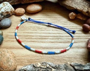 May include: A beaded bracelet with red, white, and blue beads, strung on a blue cord. The bracelet is on a wooden surface, surrounded by rocks and wood pieces.