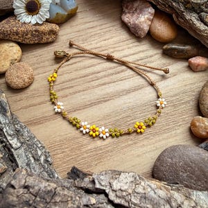 May include: A beaded bracelet featuring a daisy design with yellow, white, and green beads. The bracelet is strung on a brown cord and is adjustable. The bracelet is displayed on a wooden surface with decorative stones and bark.