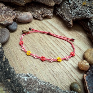 May include: A pink beaded bracelet with small yellow and red flower accents. The bracelet is made with a cord and has a sliding knot closure.