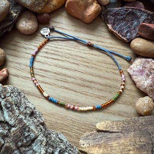 May include: A beaded bracelet with a blue cord and a variety of colorful beads. The beads include shades of brown, blue, green, pink, and cream. The bracelet is set against a wooden background with rocks and wood pieces.