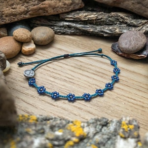 May include: A blue flower bracelet with a teal cord. The bracelet features small, dark blue flower beads with silver accents. A small, round silver charm is attached to the cord. The bracelet is displayed on a wooden surface with rocks and wood pieces in the background.