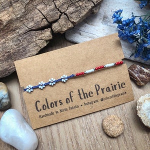 May include: A handmade beaded bracelet featuring white daisy-like flowers and red, white, and silver beads. The bracelet is displayed on a brown card with the text "Colors of the Prairie" and "Handmade in North Dakota."
