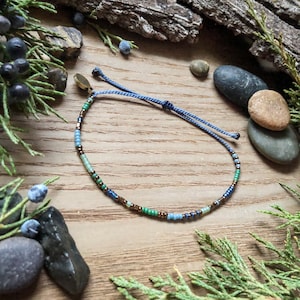 May include: A delicate blue, green, and brown beaded bracelet with a sliding knot closure. The bracelet is photographed on a wooden surface with small stones and greenery.