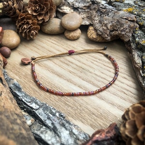May include: A delicate bracelet with small, multi-colored beads in shades of brown, red, and purple. The bracelet is made with a brown cord and a small copper charm.
