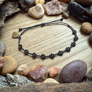 May include: A black cord bracelet with small, dark flower-shaped beads. The bracelet is adorned with a small, gold-colored tag that reads "Colors of the Prairie". The bracelet is surrounded by various rocks and wood.