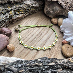 May include: A green string bracelet with white daisy beads and a gold clasp. The bracelet is on a wooden surface with rocks and a white flower.