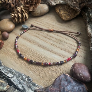 May include: A delicate beaded bracelet with a brown cord closure. The bracelet features small, colorful beads in shades of red, orange, and brown.