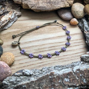 May include: A bracelet made of olive green cord featuring small purple beaded flowers arranged in a row. The bracelet includes an adjustable sliding knot closure and a small, round, metallic charm with an embossed tree design attached to the cord.