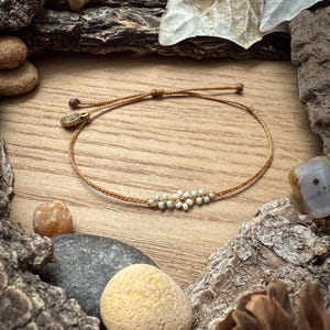 May include: A delicate, handmade bracelet featuring a series of small, light blue and white beads arranged in a floral pattern. The bracelet is strung on a thin, tan cord and has a small, gold-colored charm. The bracelet is displayed on a wooden surface.