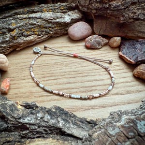 May include: A delicate beaded bracelet with a mix of white, light brown, and pink beads. The bracelet is adjustable with a sliding knot and a small charm.