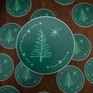 May include: A green circular sticker with a white outline features a stylized green pine tree with a sun above it. The tree is surrounded by a circle of white dots and triangles.