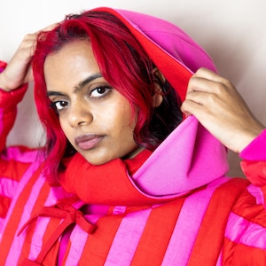 May include: A red and pink striped hooded jacket with a high collar and red ribbon ties. The hood is a contrasting pink. The person wearing the jacket has red hair. The jacket is a bold fashion statement.