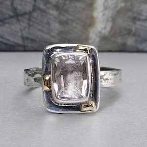 May include: A silver ring with a rectangular, light pink gemstone set in a silver frame. The ring has gold accents at each corner of the gemstone and a textured band.