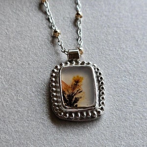 May include: A silver necklace with a square pendant featuring a natural, tree-like pattern in shades of yellow, brown, and black. The pendant is framed with a beaded border. The chain has small gold-colored beads.
